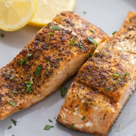 A white plate with seasoned air fryer salmon garnished with two slices of lemon on the side.