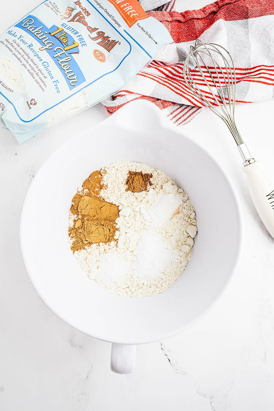 Overhead photo of dry ingredients in a white bowl. A bag of Bob's Red Mill 1 to 1 Baking Flour and a whisk are laying off to the side.