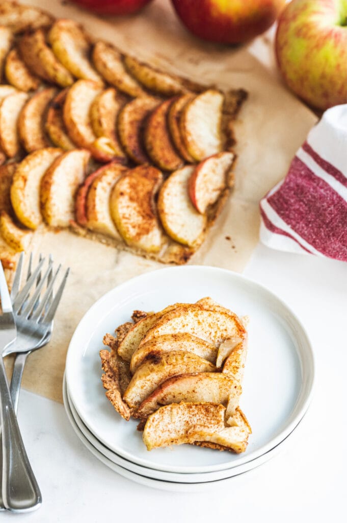 A piece of gluten free apple tart on a stack of white plates with two fork on the side. The rest of the apple tart can be seen in the background.