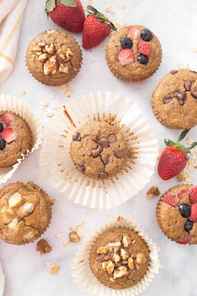 Many blender muffins with various toppings like nuts and fruit among whole strawberries, oats, and walnuts.