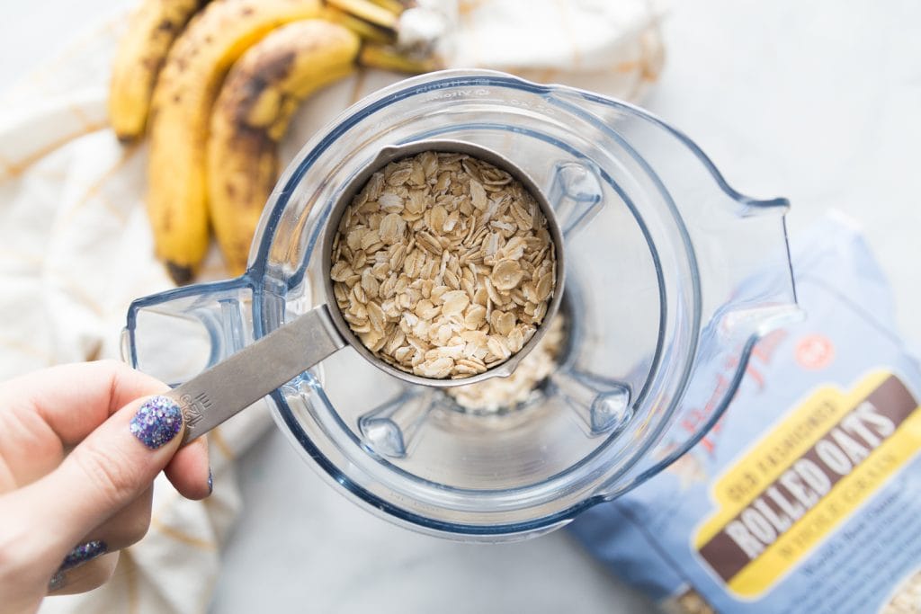 A cup of Bob's Red Mills Gluten Free Old Fashioned Oats over a blender with bananas sitting nearby. 