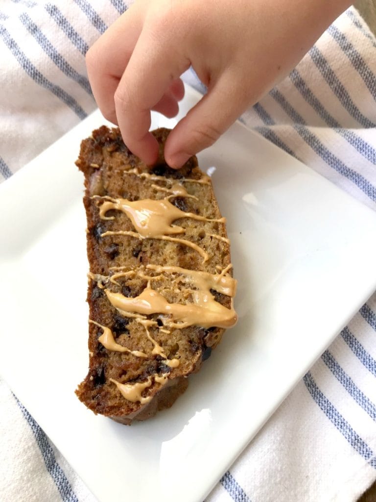 Overhead photo of a child's hand sneaking a chocolate chip from a slice of gluten free banana bread drizzled with melted peanut butter.