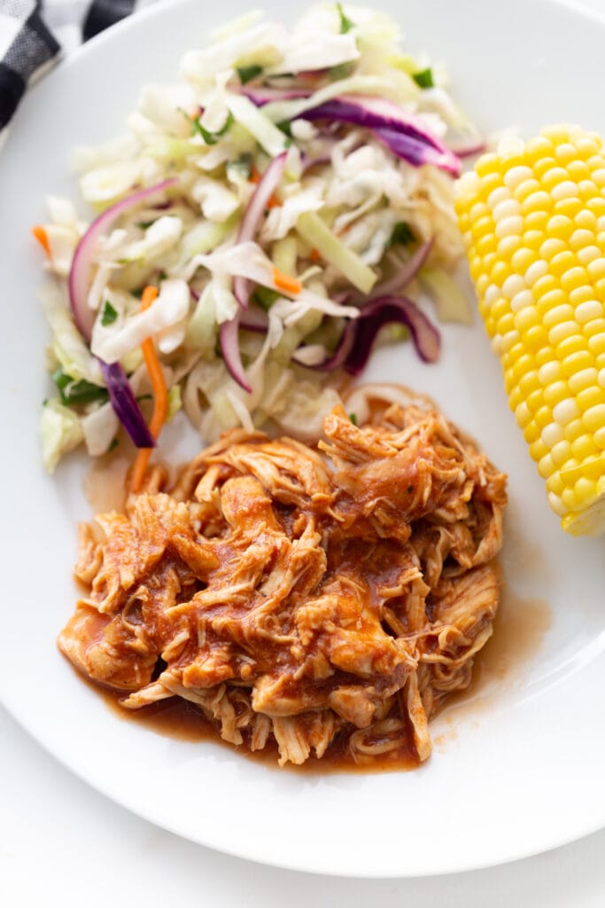 Close up photo of a white plate filled with pulled BBQ chicken, cilantro lime slaw, and corn on the cob.