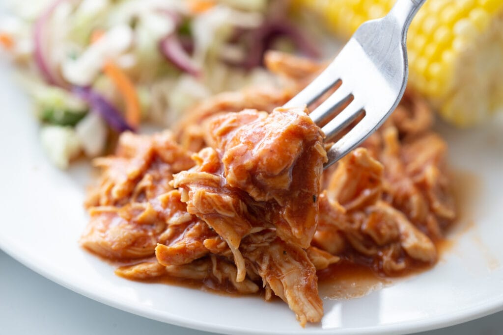 Close up photo of a fork full of juicy pulled BBQ chicken. In the background, out of focus, are homemade cole slaw and corn on the cob.