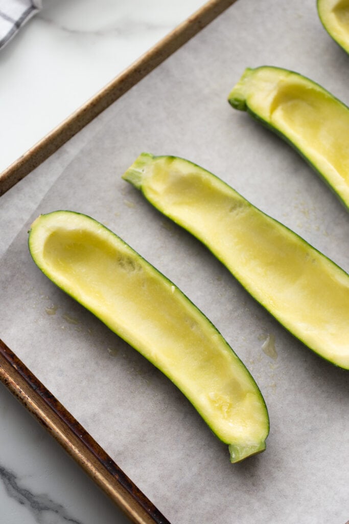 Sheet pan lined with parchment paper with halved zucchinis. The zucchini flesh has been taken out so they are ready to be stuffed.