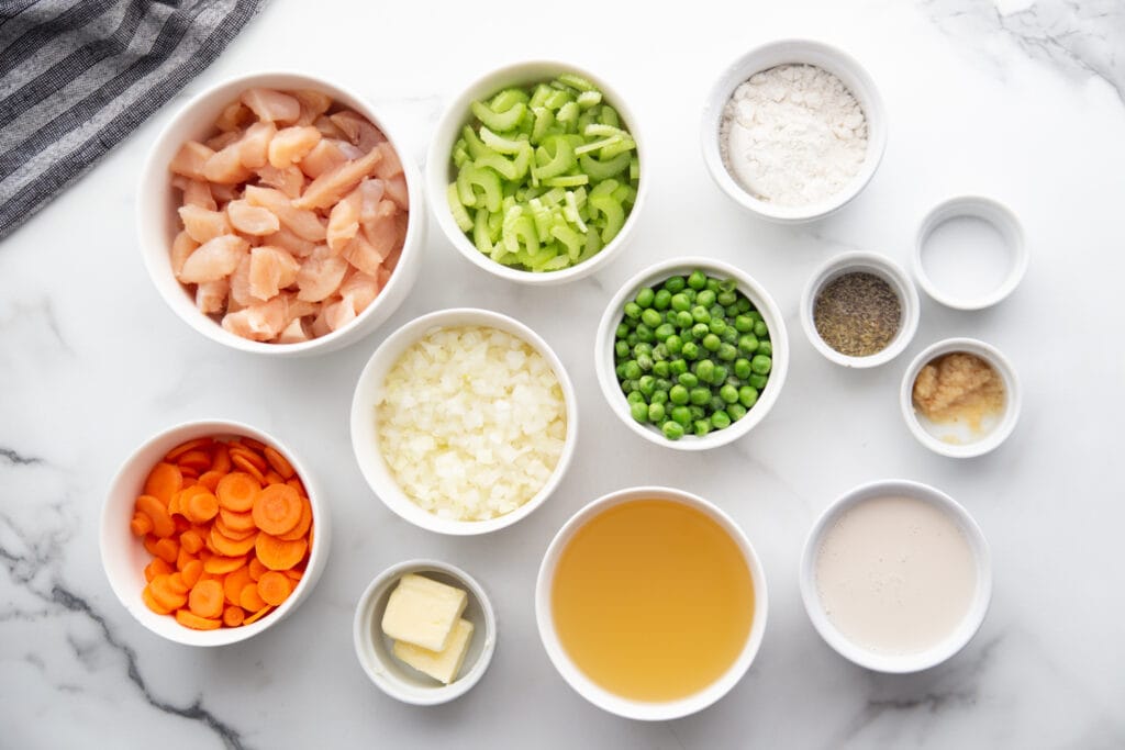 Ingredients for homemade chicken pot pie in white bowls on a white and grey marble countertop. 