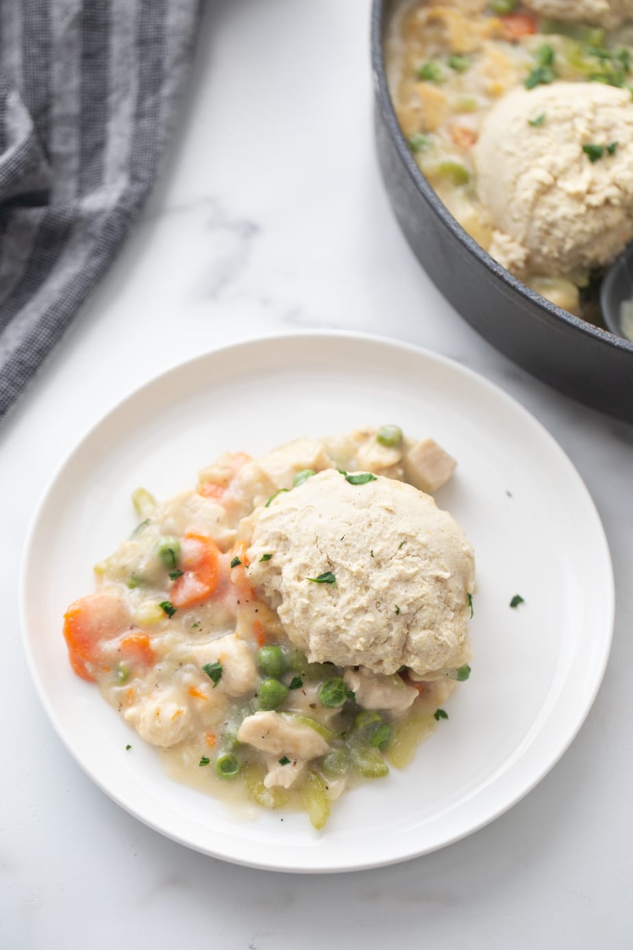 Skillet chicken pot pie with gluten free biscuit topping is piled on a white plate on a white and gray marble table. The skillet is in the upper right hand corner of the shot. 