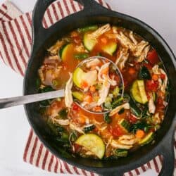 A cast iron pot of Chicken Vegetable Soup on a red and white striped kitchen towel with a small bowl of parmesan cheese off to the side.