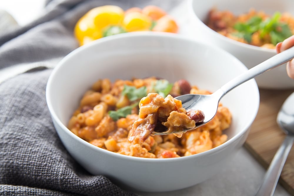 White bowl with healthy instant pot chili mac topped with cilantro. A hand is holding a spoon and about to take a bite. There is a second bowl seen in the background and some bell peppers sitting nearby. 