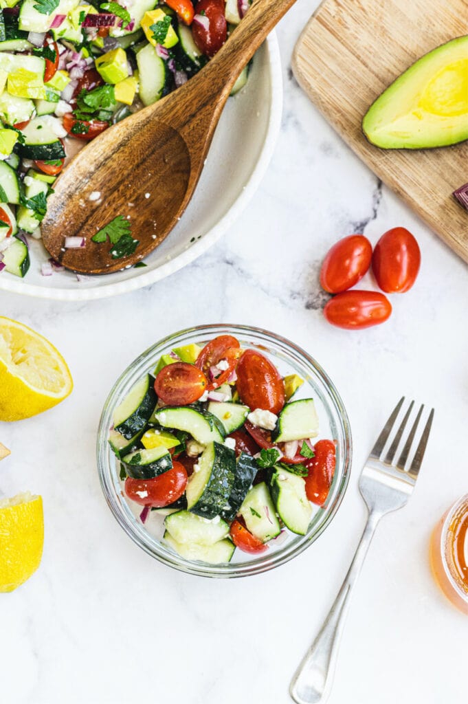 Cucumber tomato feta salad in a small glass bowl with a large serving bowl full of salad off to the side. 