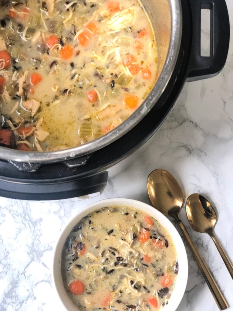 Overhead photo of cooked Instant Pot Chicken and Wild Rice Soup dished up in a white bowl with spoons next to it. The rest of the batch of soup remains in the Instant Pot.