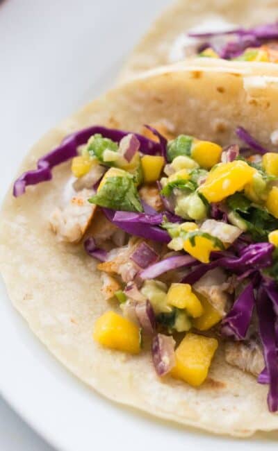 Two Fish Tacos with Mango Salsa and red cabbage on corn tortillas on a white plate.