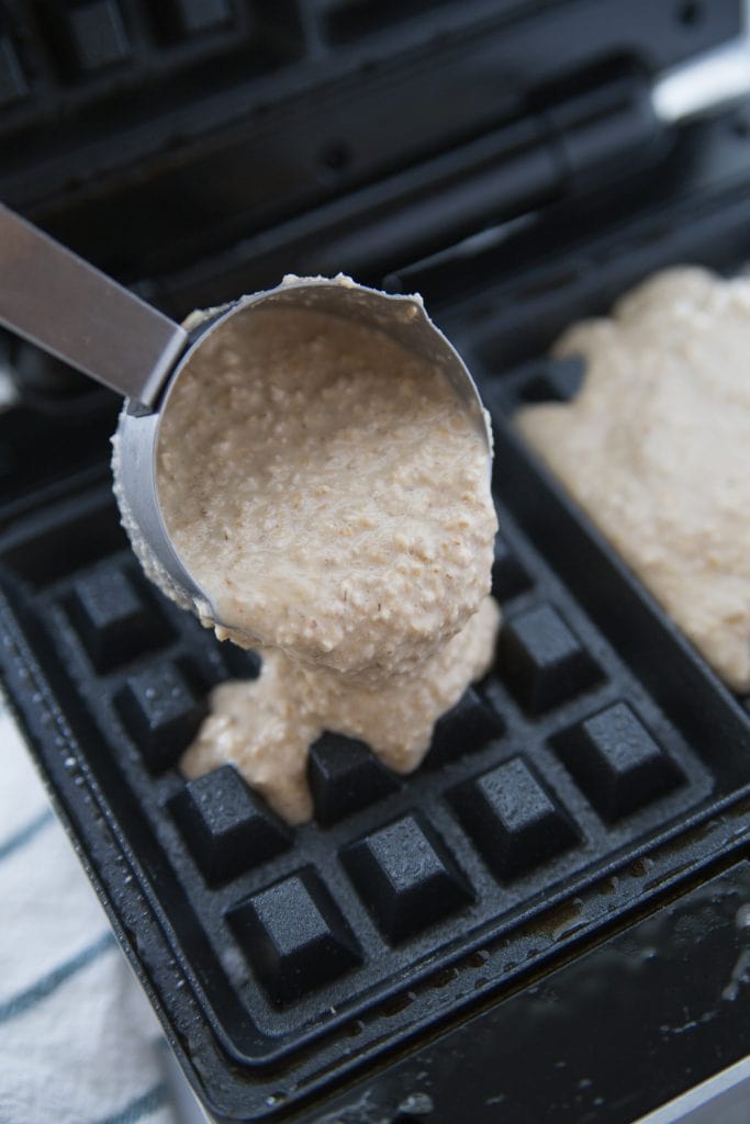 Waffle batter being poured into a hot waffle maker using a measuring cup