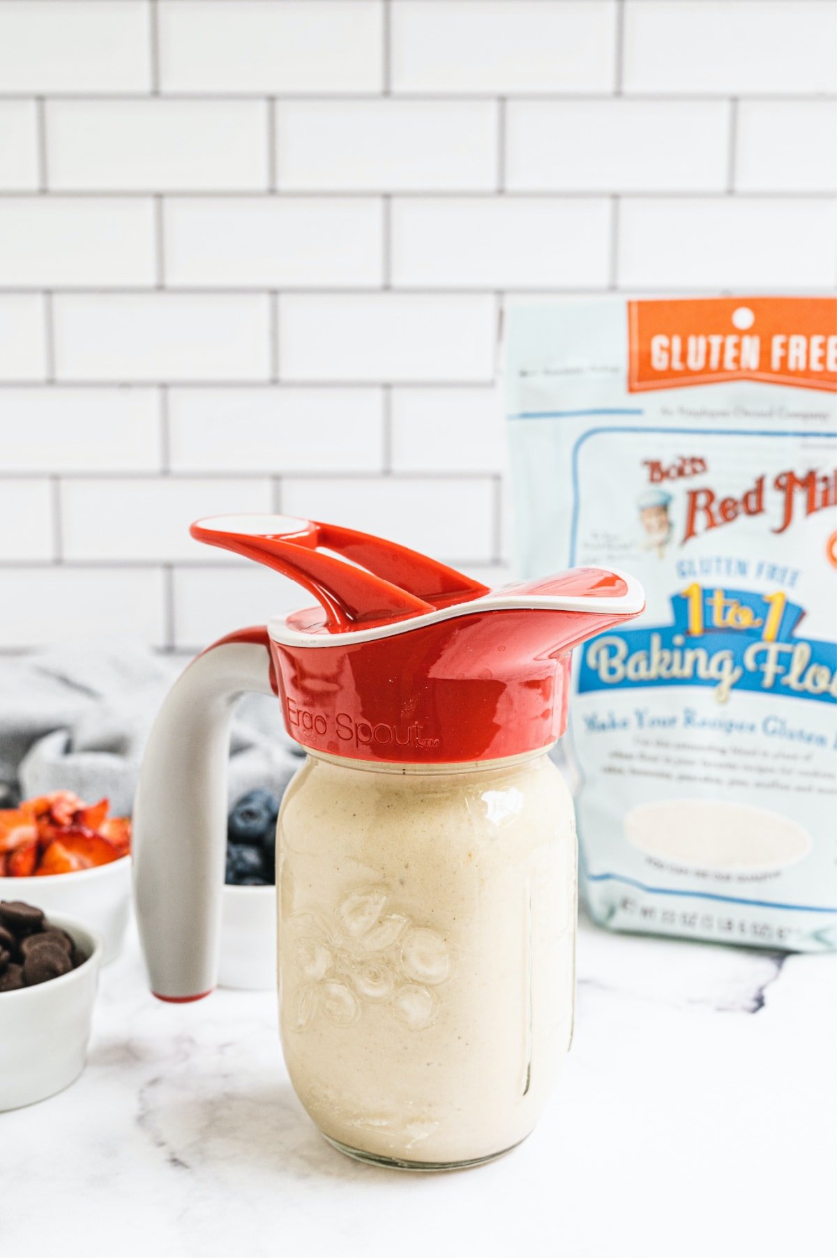 Side view of a mason jar filled with pancake batter with a red Ergo spout lid. In the background is gluten free flour and pancake mix ins.