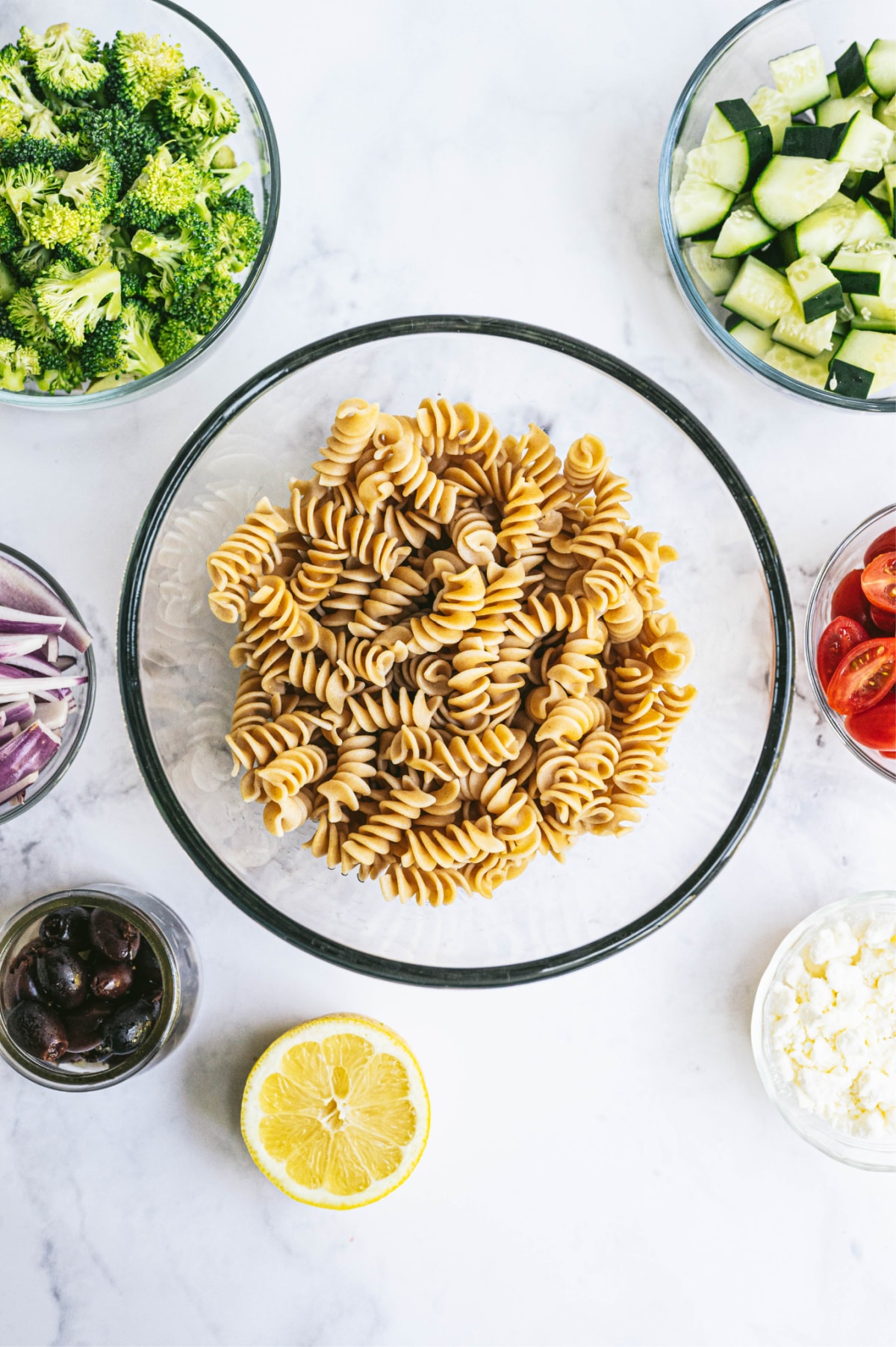 Individual bowls of varying sizes containing ingredients showing how to make pasta salad.