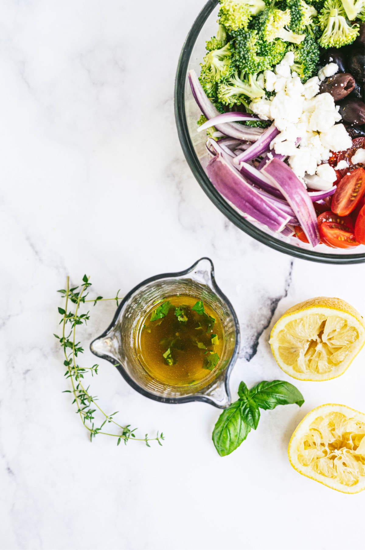 Overhead photo of glass container full of dressing, squeezed lemon halves, and a large glass bowl full of veggies and feta cheese.