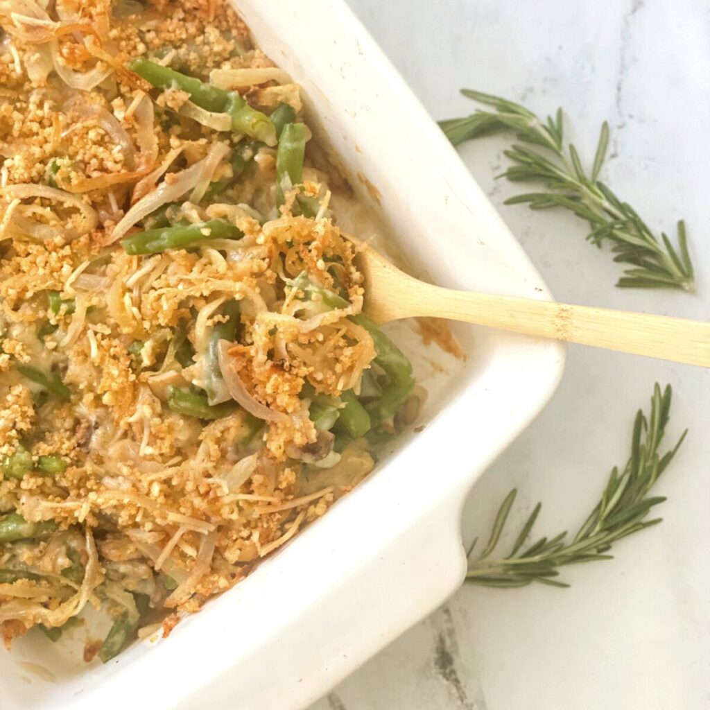 Overhead image: green bean casserole with crispy onion topping in a white casserole dish with a wooden spoon.