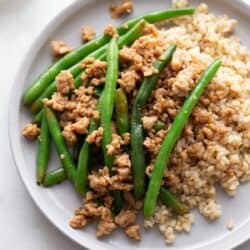 A plate of ground chicken stir fry on a bed of brown rice and stopped with steamed green beans.