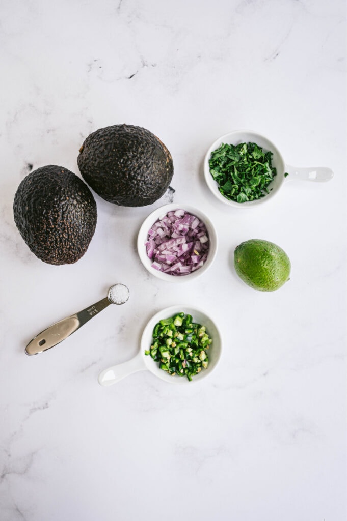 Avocado, onion, lime, salt, and cilantro sitting on a white table for guacamole 