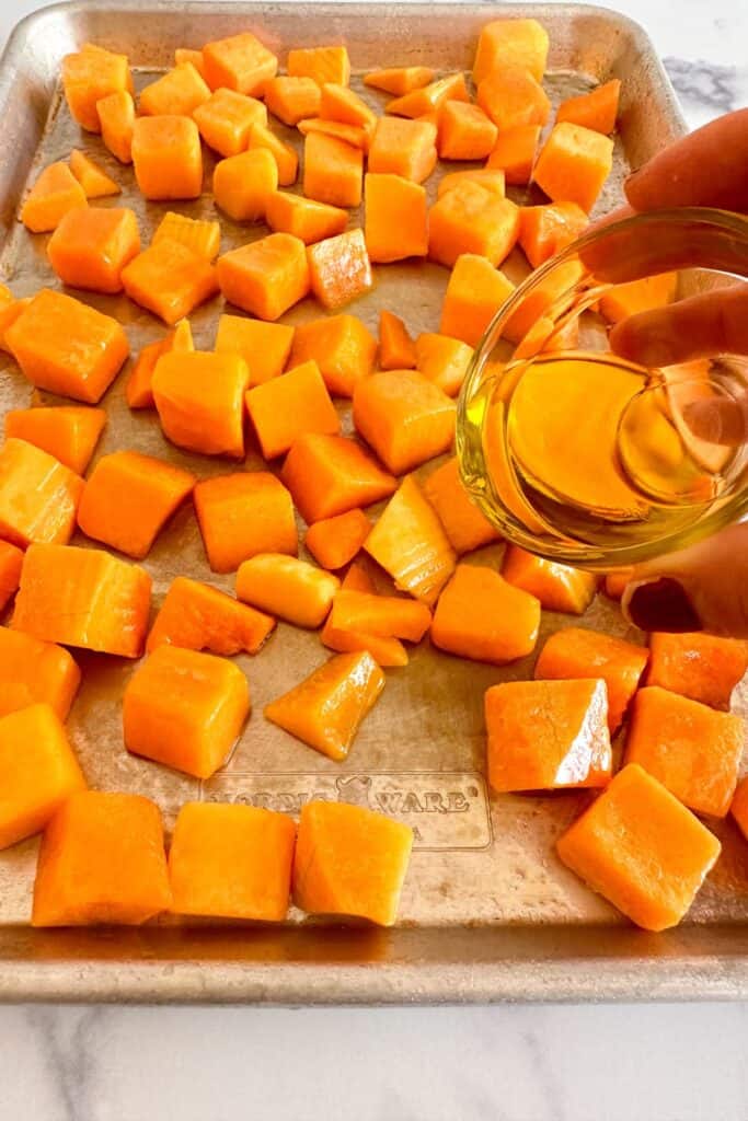 Olive oil being drizzled onto butternut squash cubes on a sheet pan.