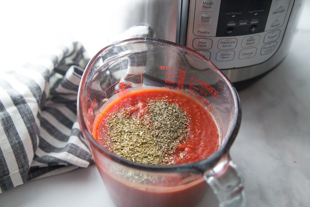 Clear glass measuring cup with tomato sauce and dried herbs in front of an Instant Pot on a marble countertop