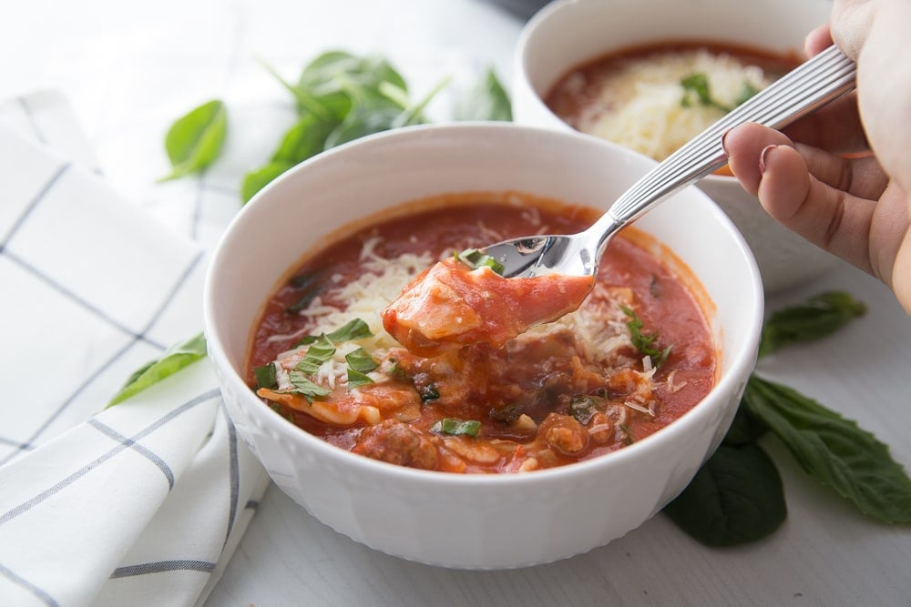 A bowl of Instant Pot Lasagna Soup sitting next to a blue and white kitchen towel. Fresh basil leaves are scattered around the table