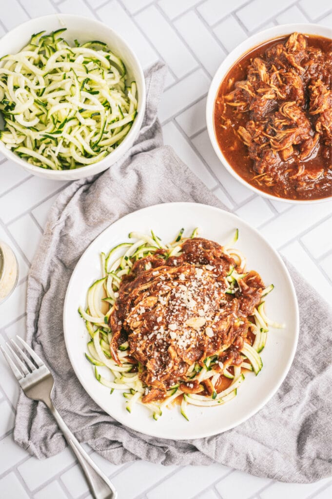 Close up overhead photo of chicken with zoodles on a white plate on a gray kitchen towel. There is a bowl of zoodles and a bowl of chicken on the side.