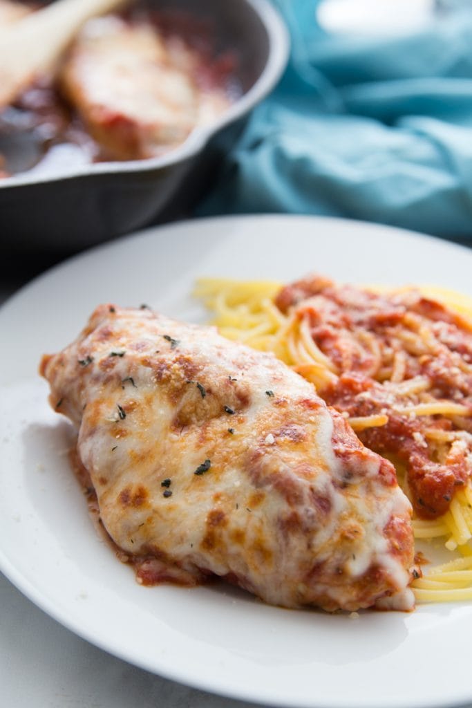 Chicken breast topped with tomato sauce, Italian spices, and melted mozzarella cheese with a side of spaghetti topped with tomato sauce on a white plate. In the background is a cast iron pan full of Italian Chicken Skillet Dinner