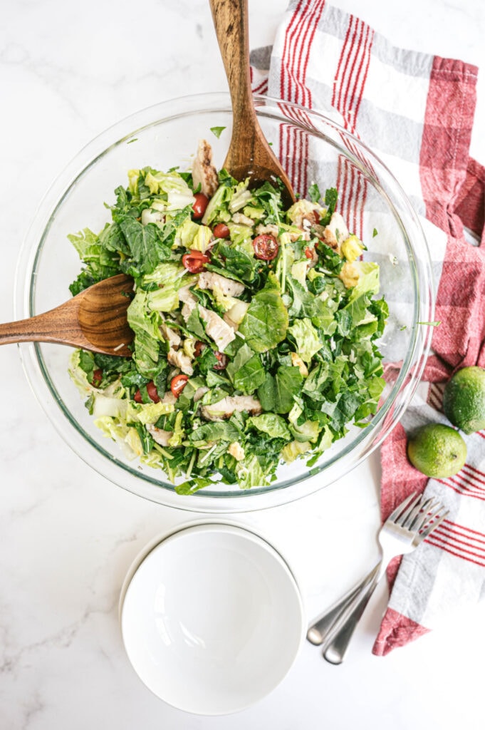 Overhead image: glass serving bowl of salad with wooden serving utensils. Empty white bowls and forks sit next to the salad.