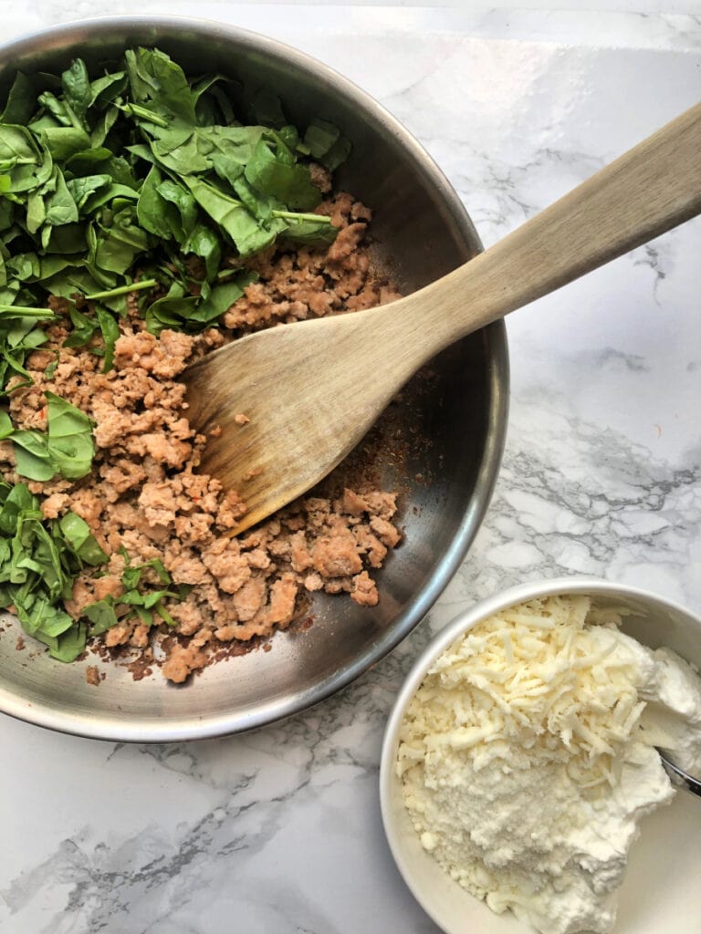 A pan with veggies and chicken sausage that will become a lasagna stuffing for Zucchini Lasagna Boats. Sitting next to the pan is a bowl of ricotta, parmesan, and mozzarella cheese. 