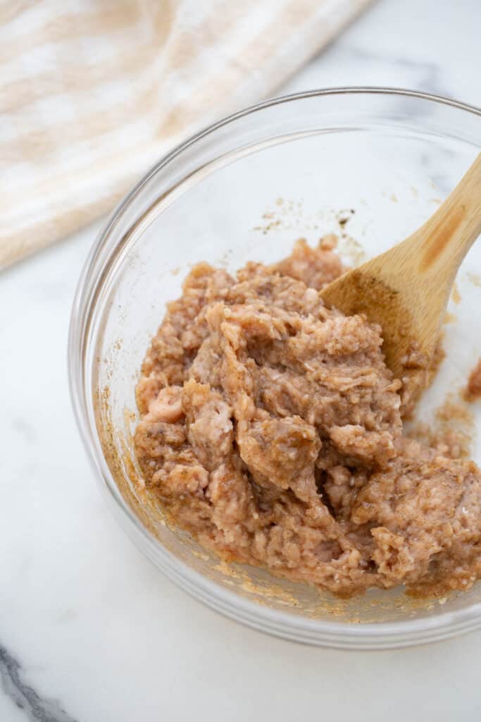 A bowl of ground chicken with a wooden spoon stirring breakfast sausage spices.