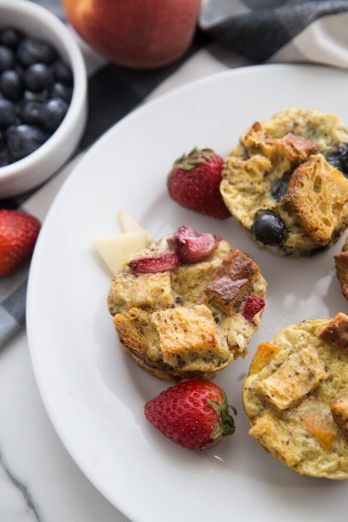 Overhead photo of Mini French Toast cups with fruit toppings and whole strawberries, on a white plate. Off to the side are whole strawberries, a peach, and a ramekin of blueberries, half out of frame.