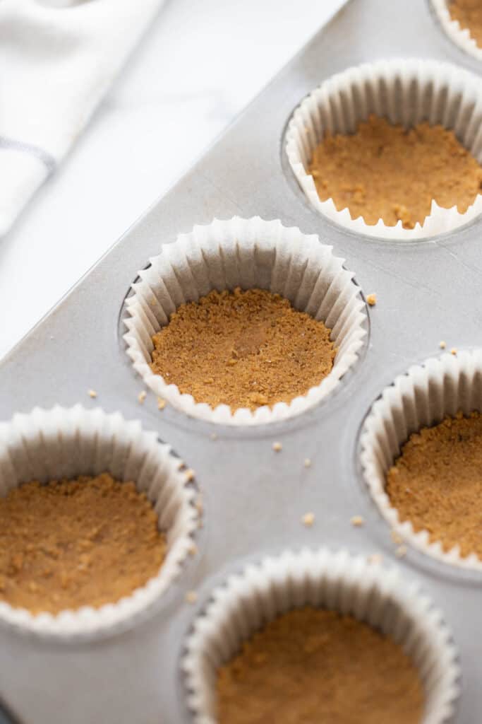 Muffin tin with parchment liners and graham cracker crust, ready for cheesecake filling.