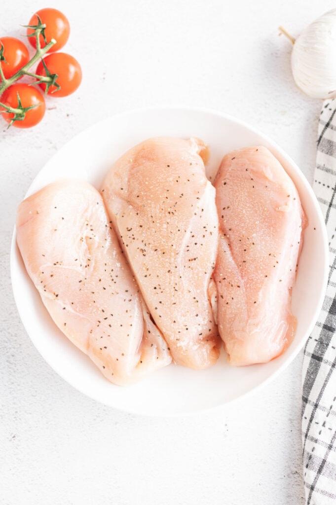 Overhead photo of three seasoned raw chicken breasts in a white bowl with whole tomatoes and garlic on the side.