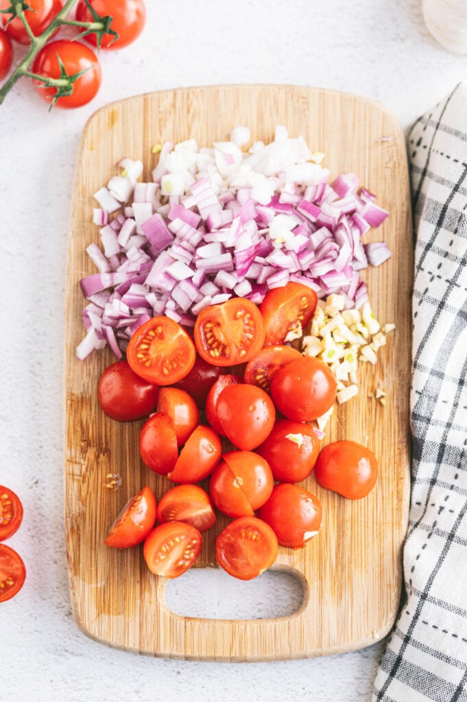 Overhead photo of halved cherry tomatoes, diced red onion, and minced garlic on a cutting board ready to make a caprese chicken recipe.