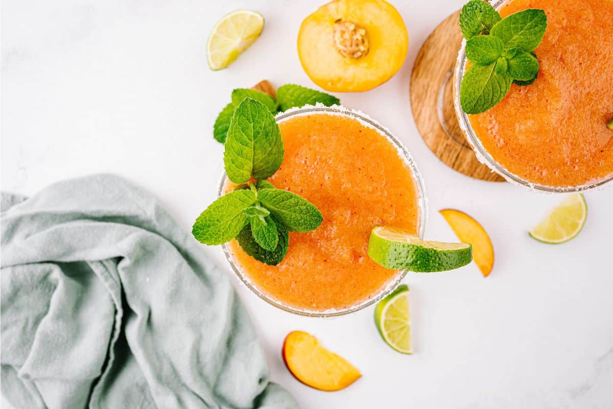 Top down shot of two frozen peach margaritas garnished with fresh mint, a lime wedge, and a salt rim.