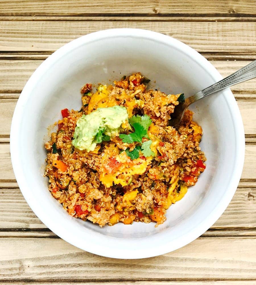 Overhead photo of Turkey Quinoa Taco Bake topped with cilantro and avocado in a white bowl with a spoon on a wooden background.