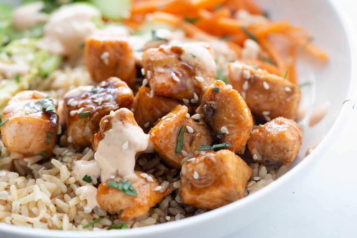 Bowl of sticky air fryer salmon bites over brown rice with spicy mayo sesame seeds and cilantro.