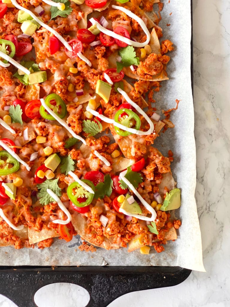 Homemade nachos topped with ground beef, cheese, jalapeno, tomatoes, corn, cilantro, avocado, and drizzled Greek yogurt on a parchment lined sheet pan.