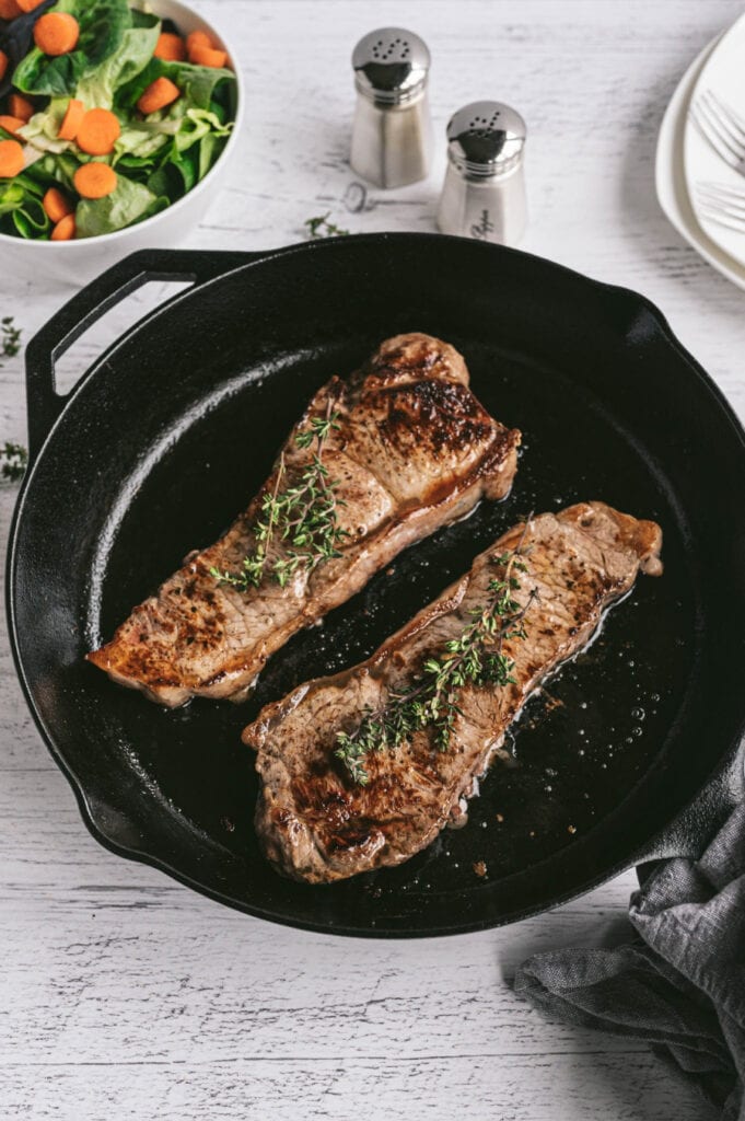 Two New York Strip Steaks grilled in a Cast Iron Skillet with salt and pepper shakers and a salad on the side.