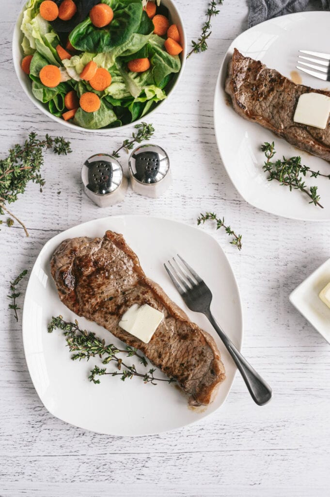 Two plates with grilled New York Strip Steak topped with a thin pad of butter and a large salad on the side.