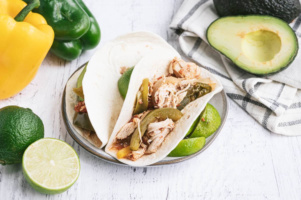 White corn tortillas with crockpot chicken fajitas inside on a plate with sliced lime.