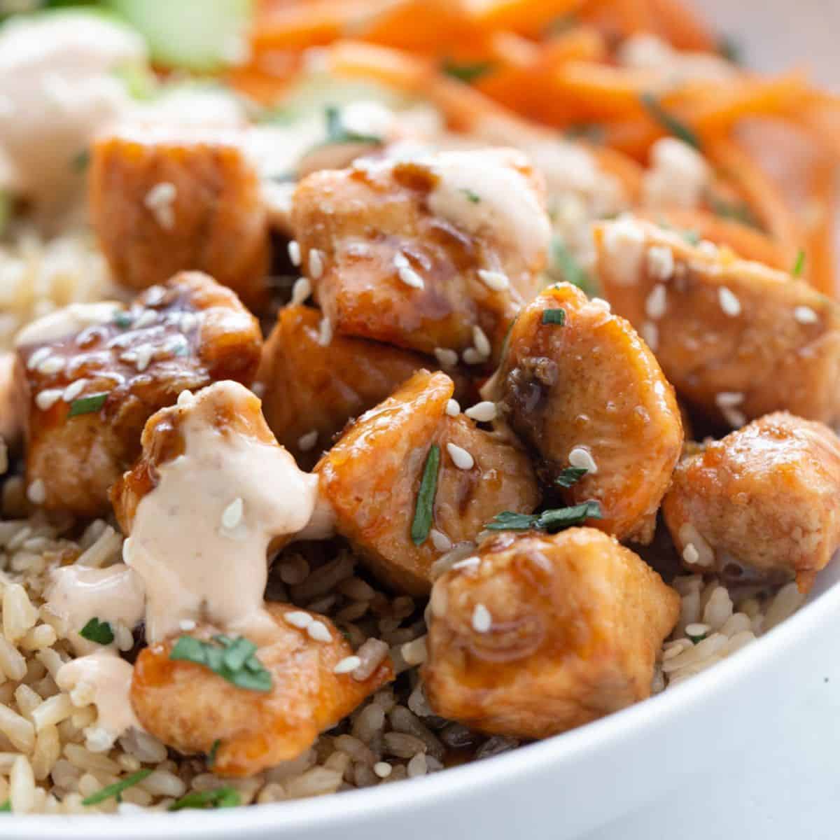 Bowl of sticky air fryer salmon bites over brown rice with spicy mayo sesame seeds and cilantro.