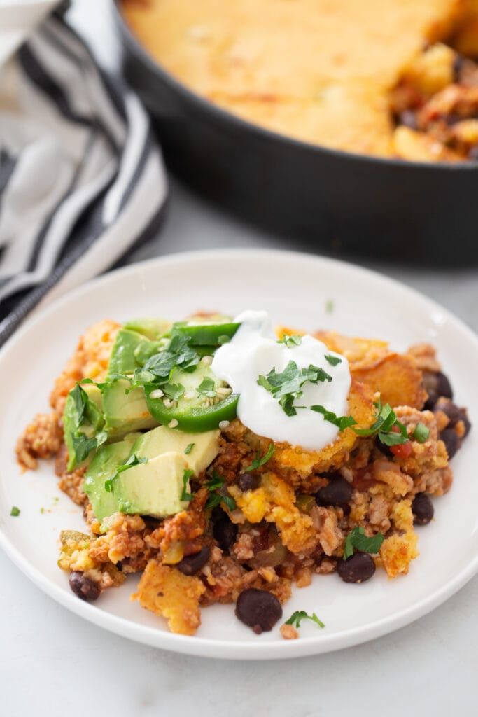 High angle photo of one pan taco chili cornbread topped with avocado, jalapenos, sour cream and cilantro, on a white plate.