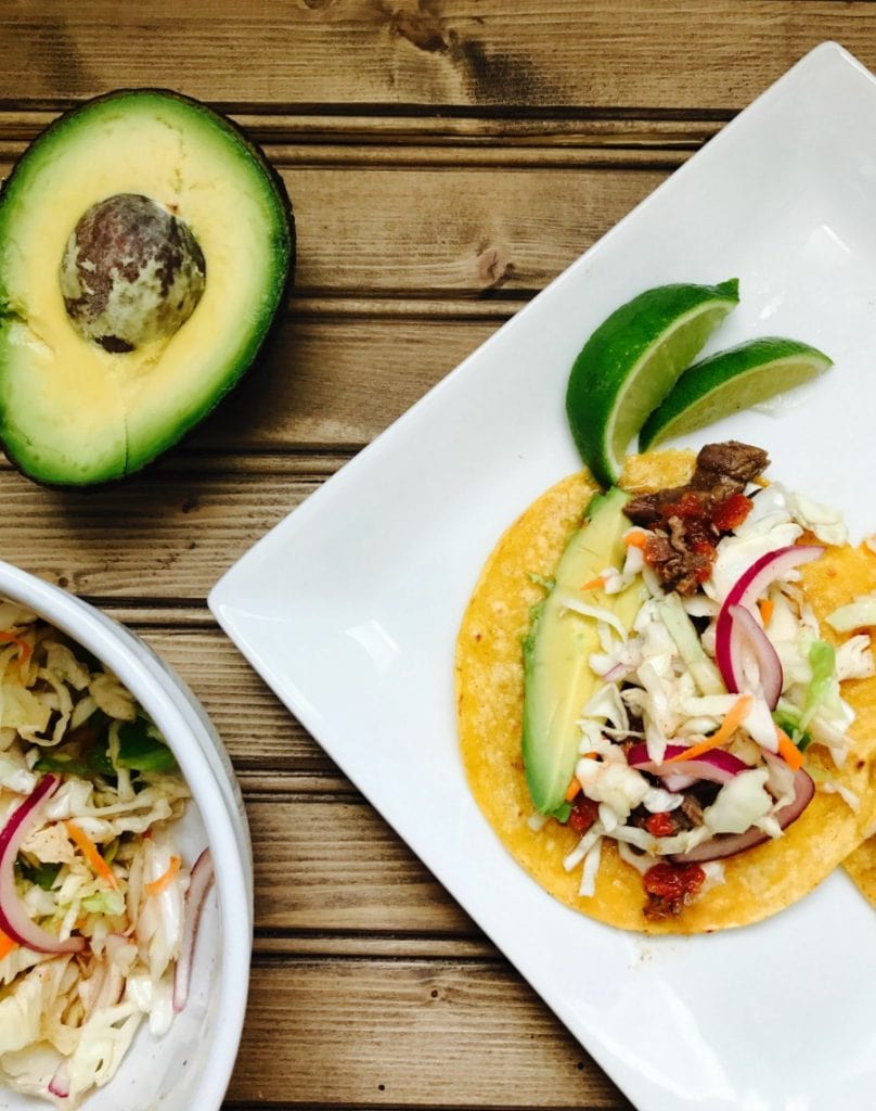 An overhead photo of an Instant Pot Flank Steak Taco laying on a white plate with limes on the side, on a wooden surface. To the side is half an avocado and a white bowl of extra cilantro lime coleslaw. In the tacos are flank steak, cilantro lime slaw, and topped with cilantro and avocado.