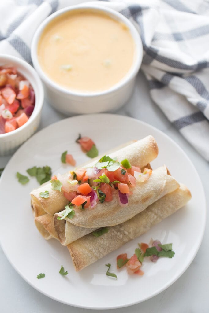 Overhead photo of a plate stacked with beef taquitos topped with homemade salsa. Ramekins filled with salsa and homemade cheese sauce are next to the plate.