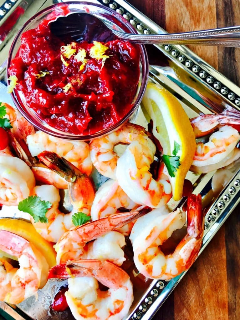 Roasted Shrimp Cocktail on a silver platter with Cranberry Horseradish Chutney in a glass bowl. Lemon wedges and cilantro for garnish.