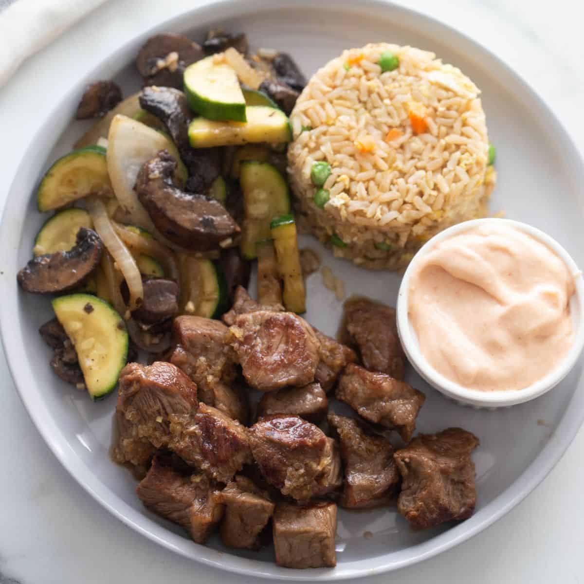 White plate with hibachi steak, hibachi veggies, yum yum sauce, and fried rice.