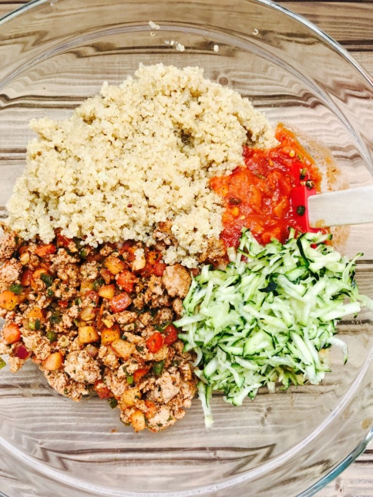 Deconstructed ingredients for a Turkey Quinoa Taco Bake- Sauteed turkey and veggies, shredded zucchini, cooked quinoa, and tomato sauce in a glass mixing bowl on a wooden surface.