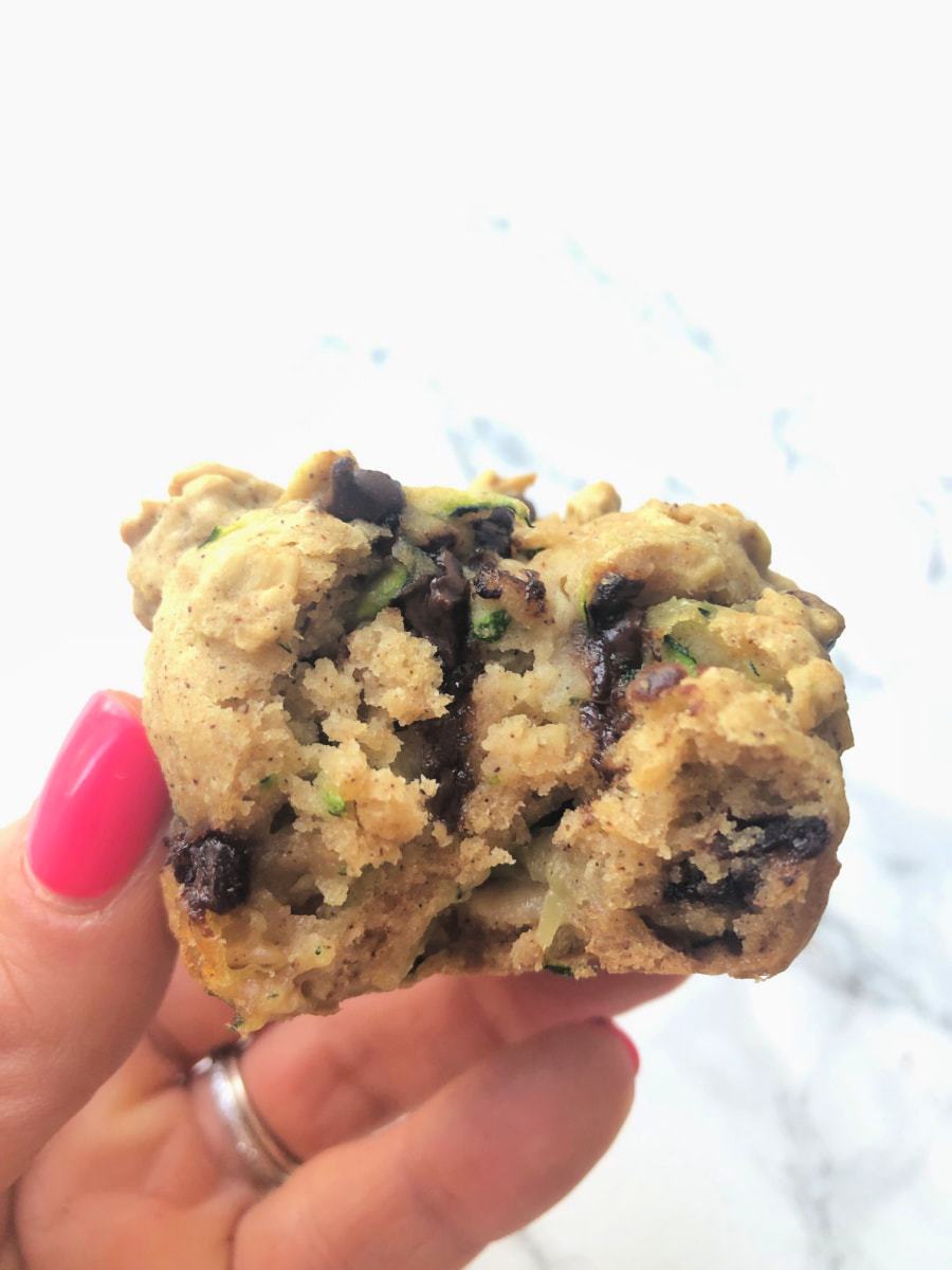 A woman's hand holding a chocolate chip zucchini muffin over a white marble background.